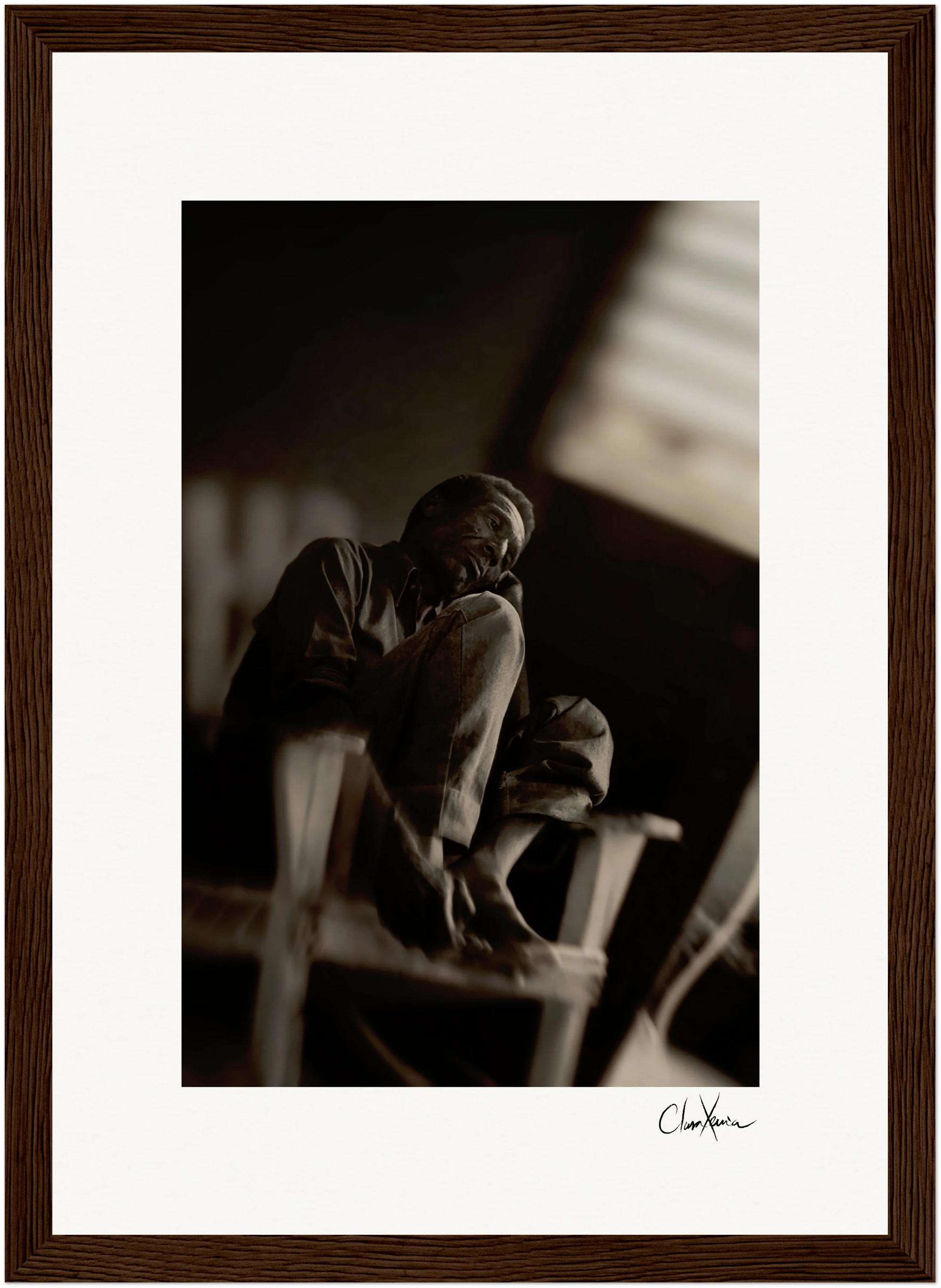 A black and white photo shows an older person sitting on a chair with one knee drawn up, gazing off to the side. Soft, diffused light from a window enhances this fine art print, framed with a dark wooden border—perfect for mindful home decor.