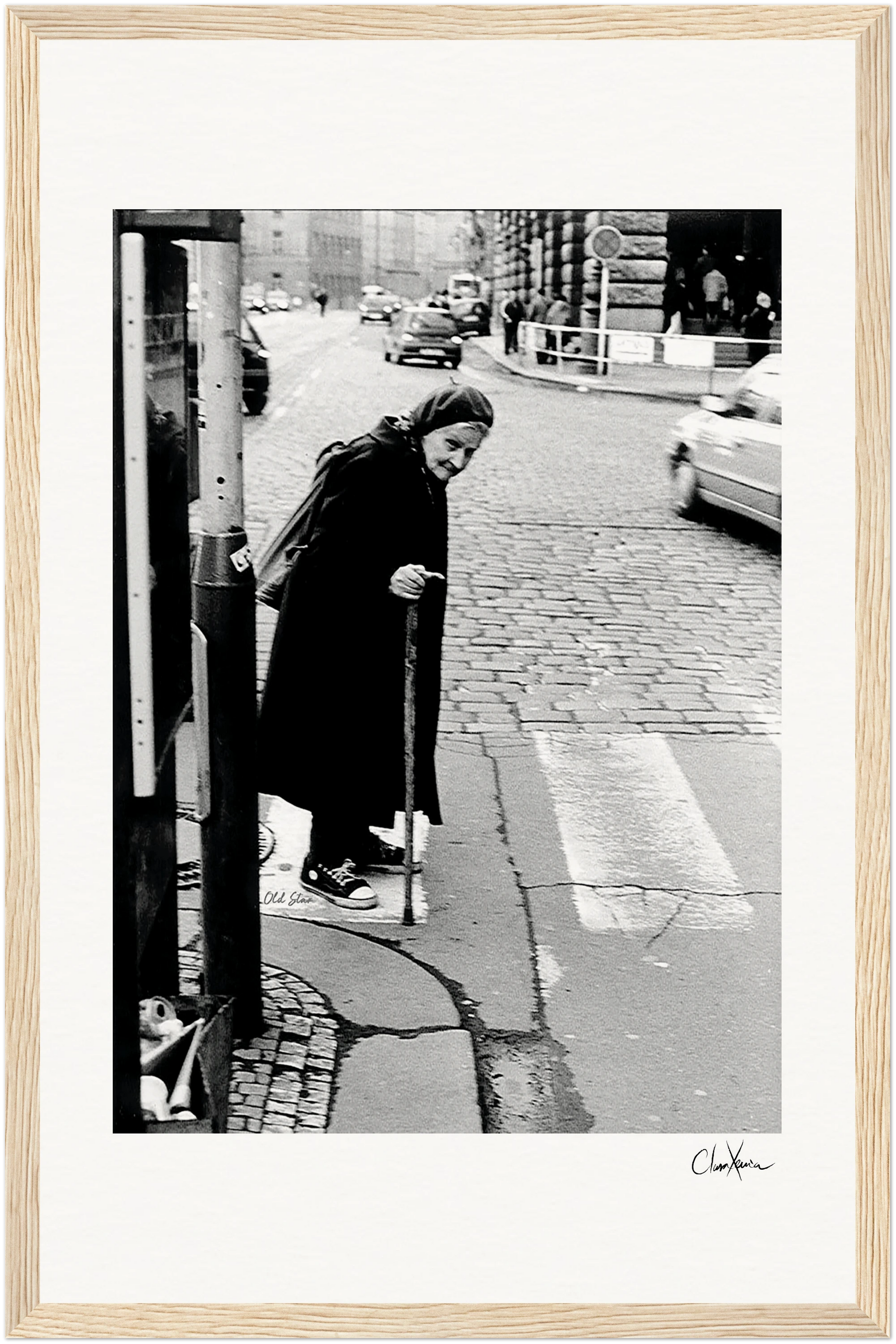 An elderly woman with a cane crosses a cobblestone street at a crosswalk in an urban setting. She wears a dark coat and scarf. Cars and buildings are visible in the background—perfect as inspirational wall art for conscious interior design.