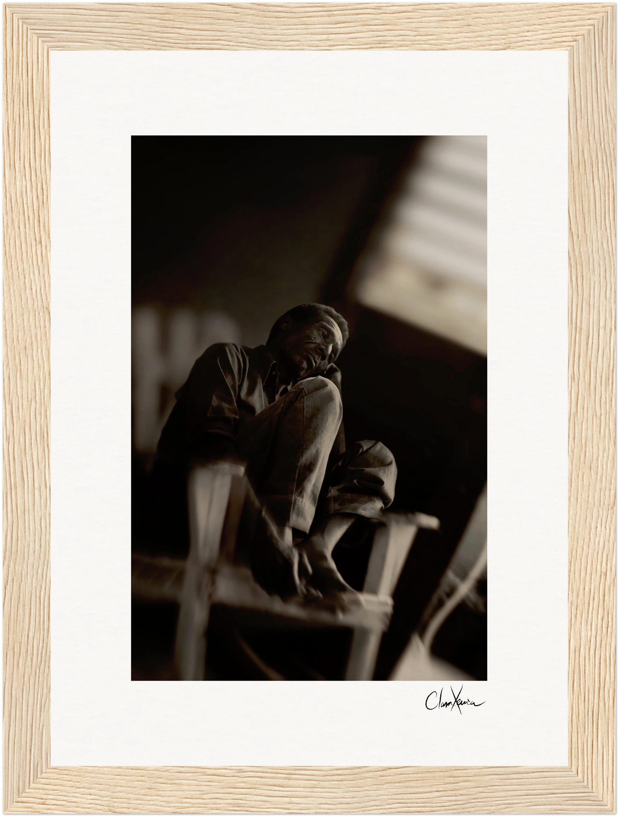 A black-and-white photo shows an older man sitting on a wooden chair with his legs up, head resting on his hand. The blurred background and dim lighting enhance this fine art print, making it perfect for mindful home decor. Displayed in a light wood frame.