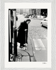 An elderly woman in a headscarf and dark clothing leans on a cane while crossing a cobblestone street at a crosswalk. This black-and-white photo makes inspirational wall art in a gallery-quality frame, perfect for mindful home decor. Cars and buildings appear behind her.
