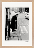 An elderly woman in a headscarf and long coat uses a cane to cross a cobblestone street at a crosswalk. Cars and buildings appear in the background. This black and white photo is presented as a fine art print in a gallery-quality frame—perfect for mindful home decor.