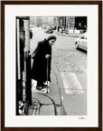 An elderly woman with a cane stands at a crosswalk on a cobblestone city street. She wears a dark coat and headscarf, gazing toward passing cars—an evocative scene perfect for mindful home decor or conscious interior design.