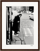 An elderly woman with a cane stands at a crosswalk on a cobblestone city street. She wears a dark coat and headscarf, gazing toward passing cars—an evocative scene perfect for mindful home decor or conscious interior design.