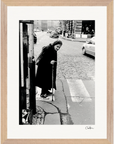 An elderly woman with a cane pauses at a crosswalk on a city street, with cars and buildings in the background. This black and white fine art print is elegantly framed in a gallery-quality frame, perfect for mindful home decor.
