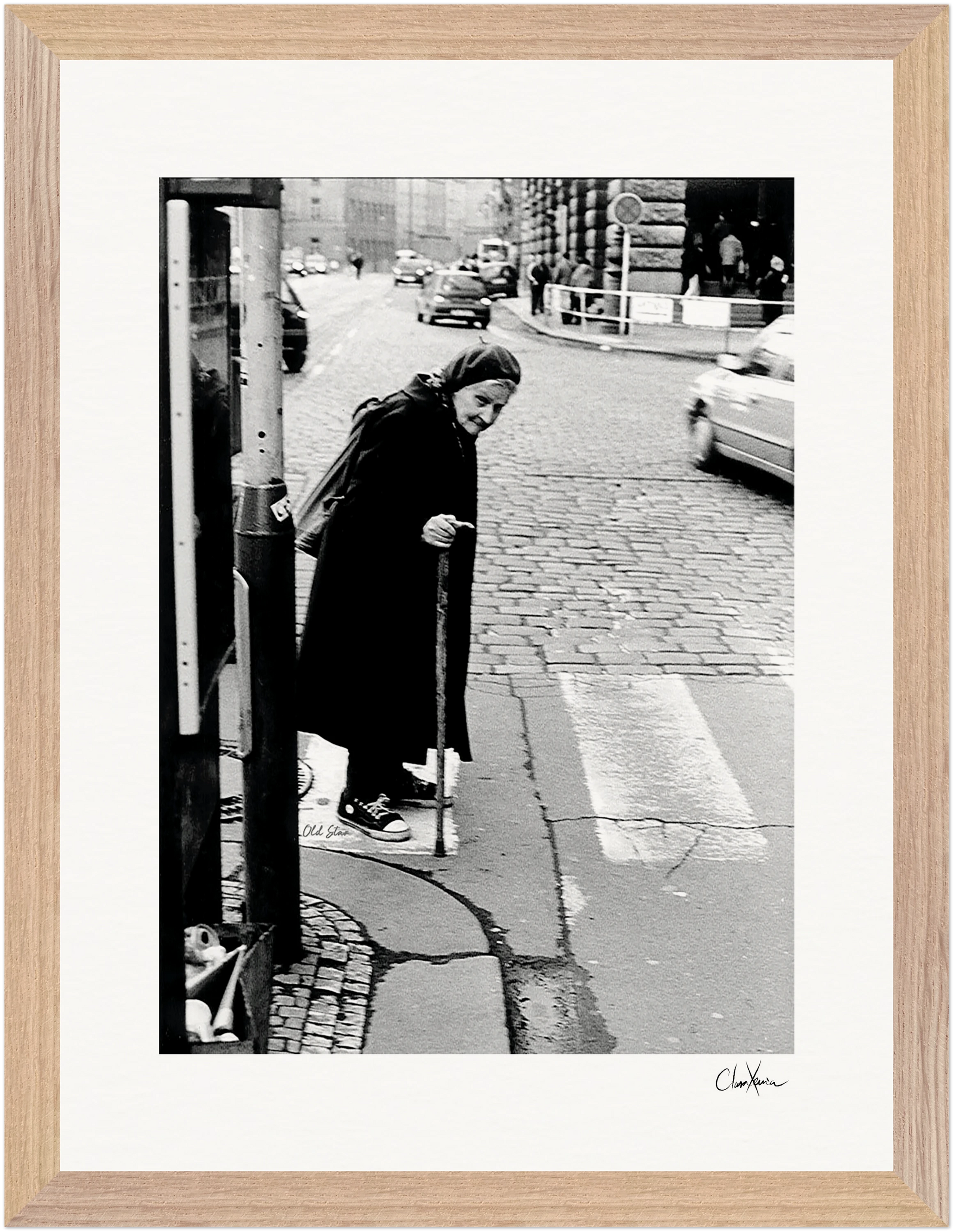 An elderly woman with a cane pauses at a crosswalk on a city street, with cars and buildings in the background. This black and white fine art print is elegantly framed in a gallery-quality frame, perfect for mindful home decor.