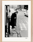 An elderly woman with a cane stands at a crosswalk on a city street, navigating a cracked sidewalk. Cars and buildings appear in the background. This black and white fine art print is beautifully presented as framed wall art.