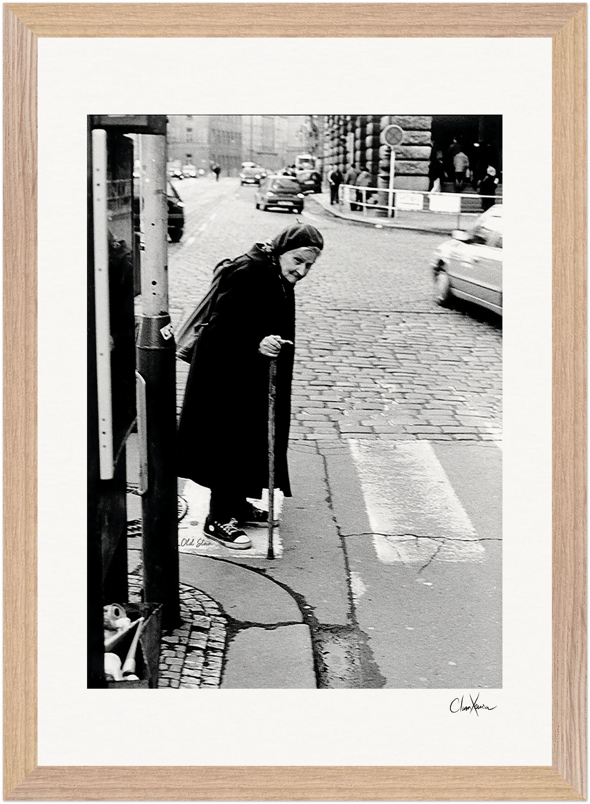 An elderly woman with a cane stands at a crosswalk on a city street, navigating a cracked sidewalk. Cars and buildings appear in the background. This black and white fine art print is beautifully presented as framed wall art.