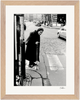 An elderly woman in dark clothing stands on a city sidewalk at a crosswalk, leaning on a cane. Cars line the cobblestone street behind her. This black and white image is presented in a gallery-quality frame, perfect for conscious interior design.