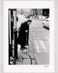 An elderly woman with a cane stands at a crosswalk on a city street, wearing a long coat and headscarf. Cars pass on the cobblestone road behind her. This scene makes for striking inspirational wall art or an elegant fine art print.