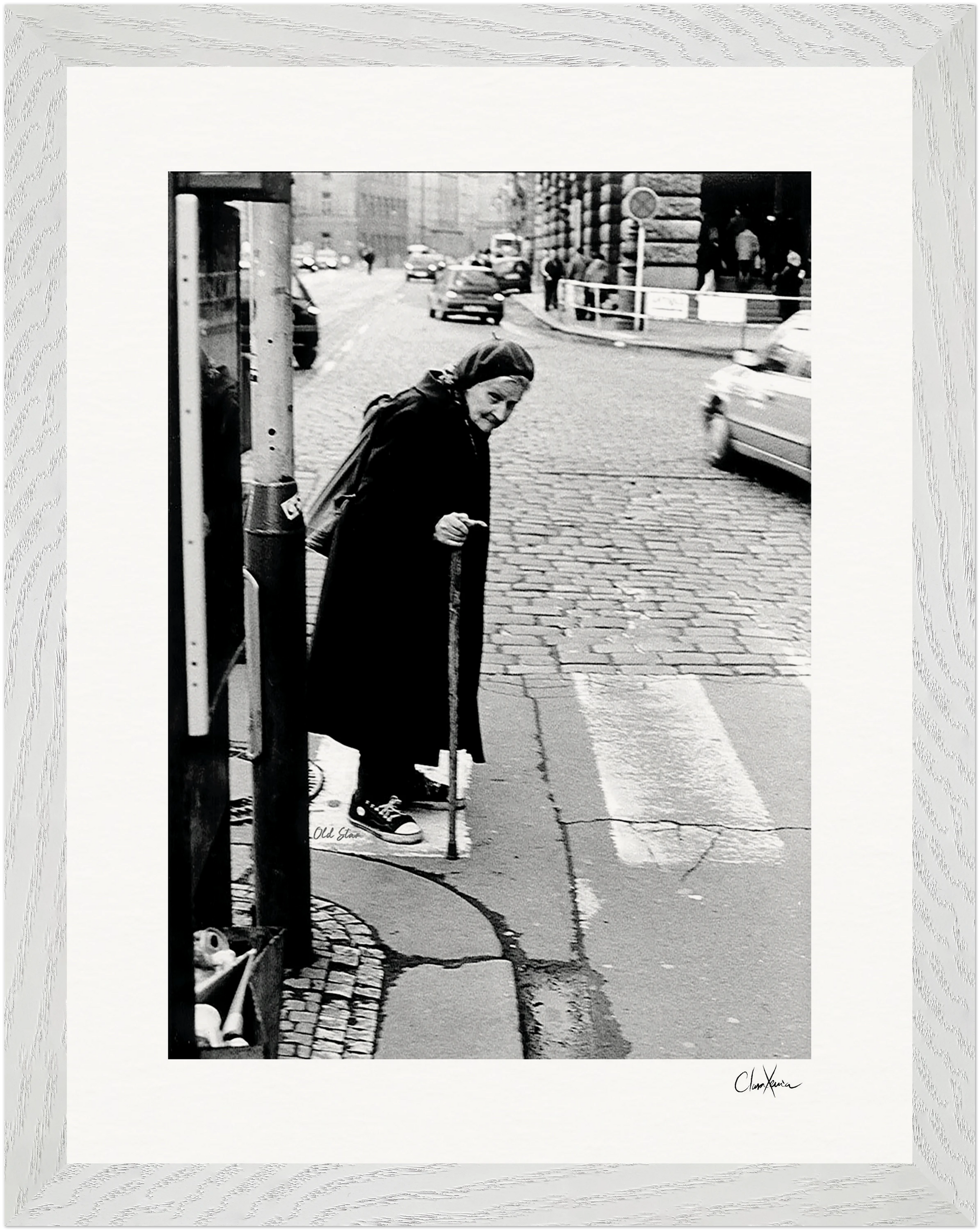 An elderly woman in a long coat and headscarf walks with a cane across a cobblestone street at a crosswalk, framed by cars and buildings—an image worthy of a gallery-quality frame, blending mindful home decor with city life.