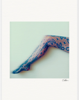A single leg extended sideways, wearing patterned, lace-like stockings with a floral design, is set against a plain, light background—perfect as a fine art print to elevate any conscious interior design.