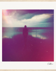 A silhouette of a person stands on the shore facing the ocean under a cloudy sky with colorful light effects and grainy texture, beautifully presented as mindful home decor in a light wooden frame.