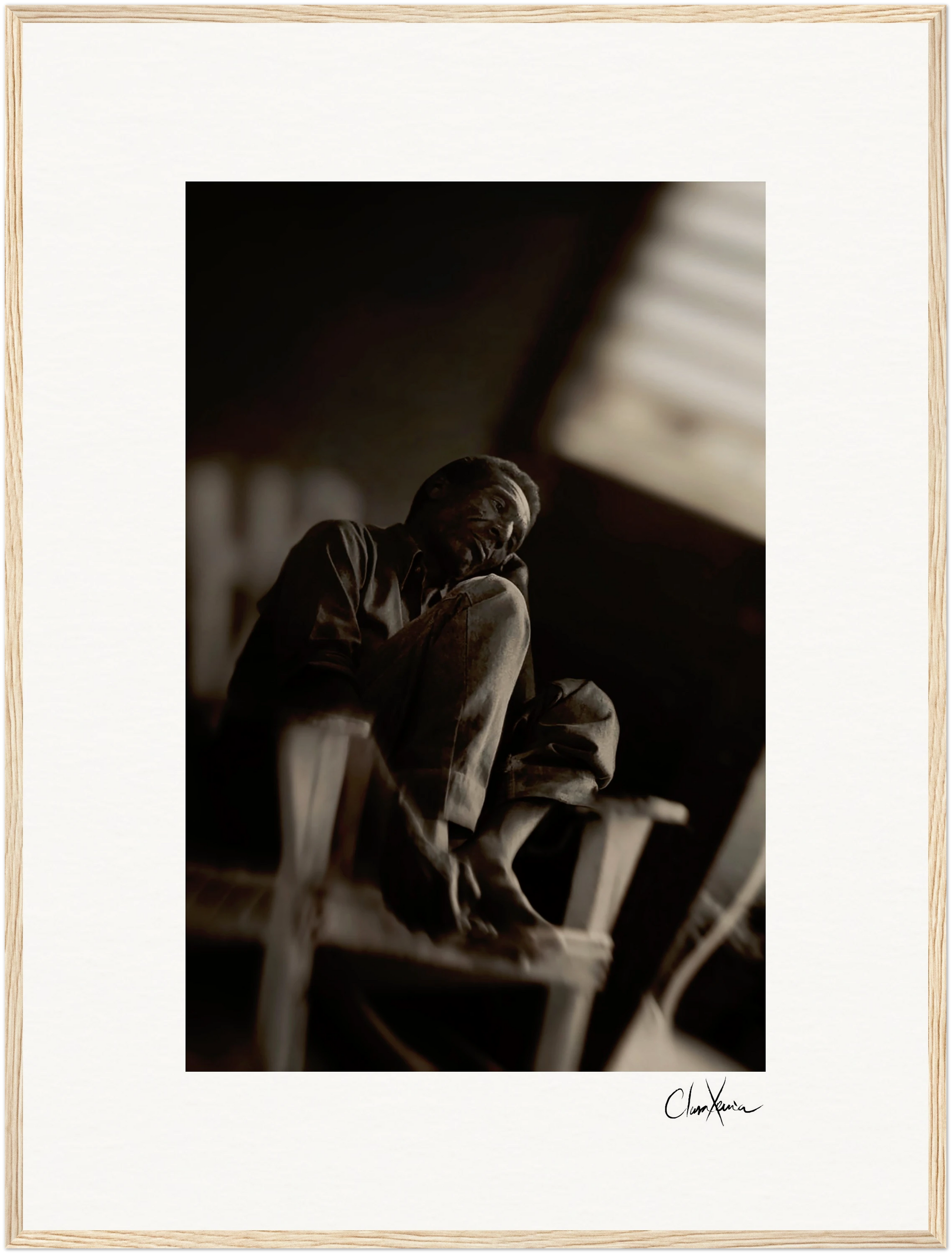 A man sits barefoot on a wooden chair indoors with one knee raised, gazing upward. The black-and-white photo features soft focus, dramatic lighting, and a visible signature—ideal as framed wall art or mindful home decor.