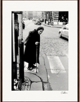 An elderly woman in a dark coat and headscarf walks with a cane on a cobblestone city street, approaching a crosswalk. Cars and buildings form the background in this black-and-white photo, perfect as inspirational wall art with a gallery-quality frame.