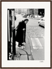 An elderly woman in a dark coat and headscarf walks with a cane on a cobblestone city street, approaching a crosswalk. Cars and buildings form the background in this black-and-white photo, perfect as inspirational wall art with a gallery-quality frame.