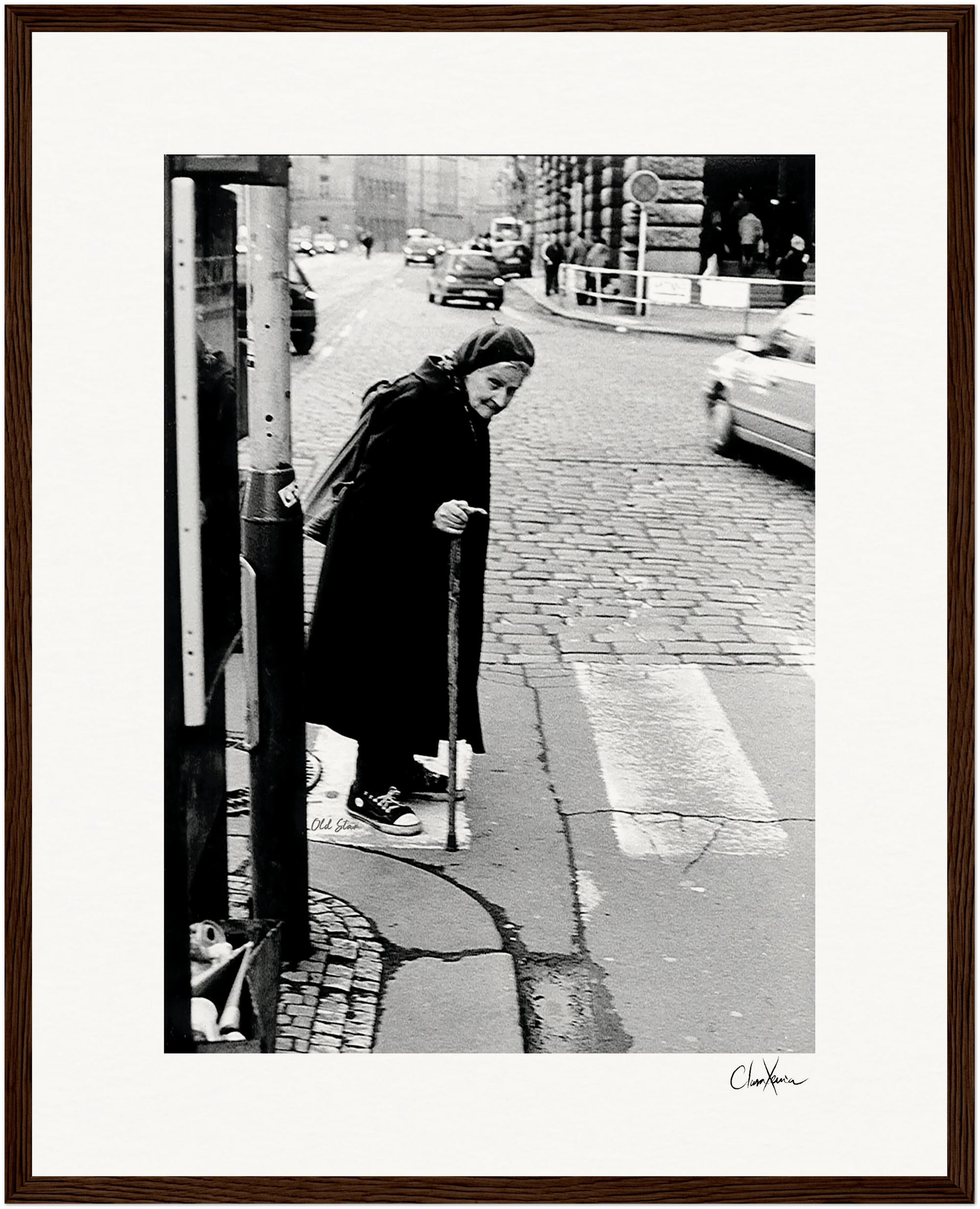 An elderly woman with a cane walks slowly across a cobblestone street at a crosswalk, framed by cars and old buildings in the background. This black and white image makes striking framed wall art for mindful home decor.