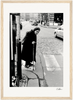 An elderly woman with a cane walks slowly across a cobblestone street at a crosswalk. She wears a long coat and headscarf. Cars and people move in the background, creating an evocative scene ideal for a fine art print or mindful home decor.