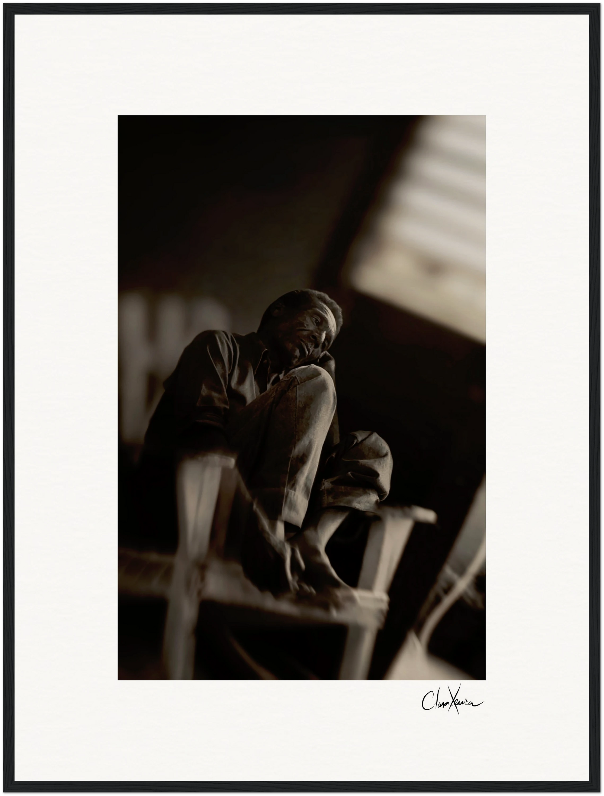 A black and white photo shows an older man sitting barefoot on a wooden chair, one knee raised. Soft light from a window highlights his thoughtful expression, making this fine art print a striking addition to conscious interior design.
