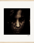 A close-up, grainy, dark-toned photograph of a person's face with long hair partially covering the features, set against a light background and presented as fine art print in a simple light wood frame—perfect for conscious interior design.