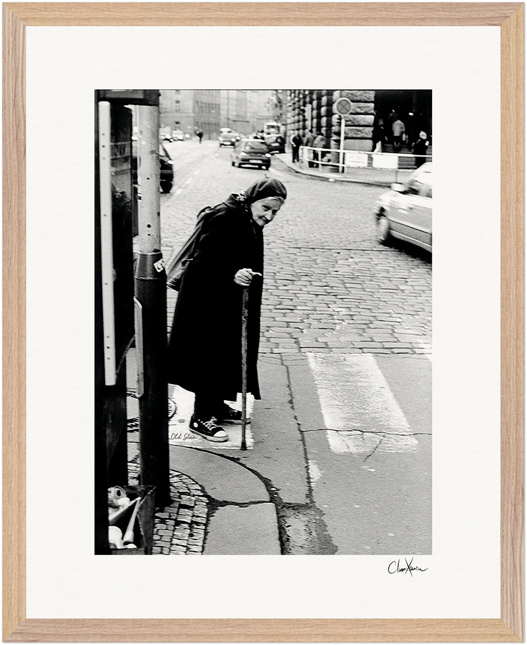 An elderly woman in dark clothing and headscarf uses a cane while crossing a cobblestone street. Cars and buildings form the background of this black-and-white fine art print, elegantly presented as framed wall art with a light wood border.