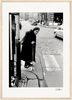 An elderly woman with a headscarf and cane stands at the edge of a cobblestone street, ready to cross. Cars and people appear in the background. This black-and-white image is framed in light wood, perfect as inspirational wall art for conscious interior design.