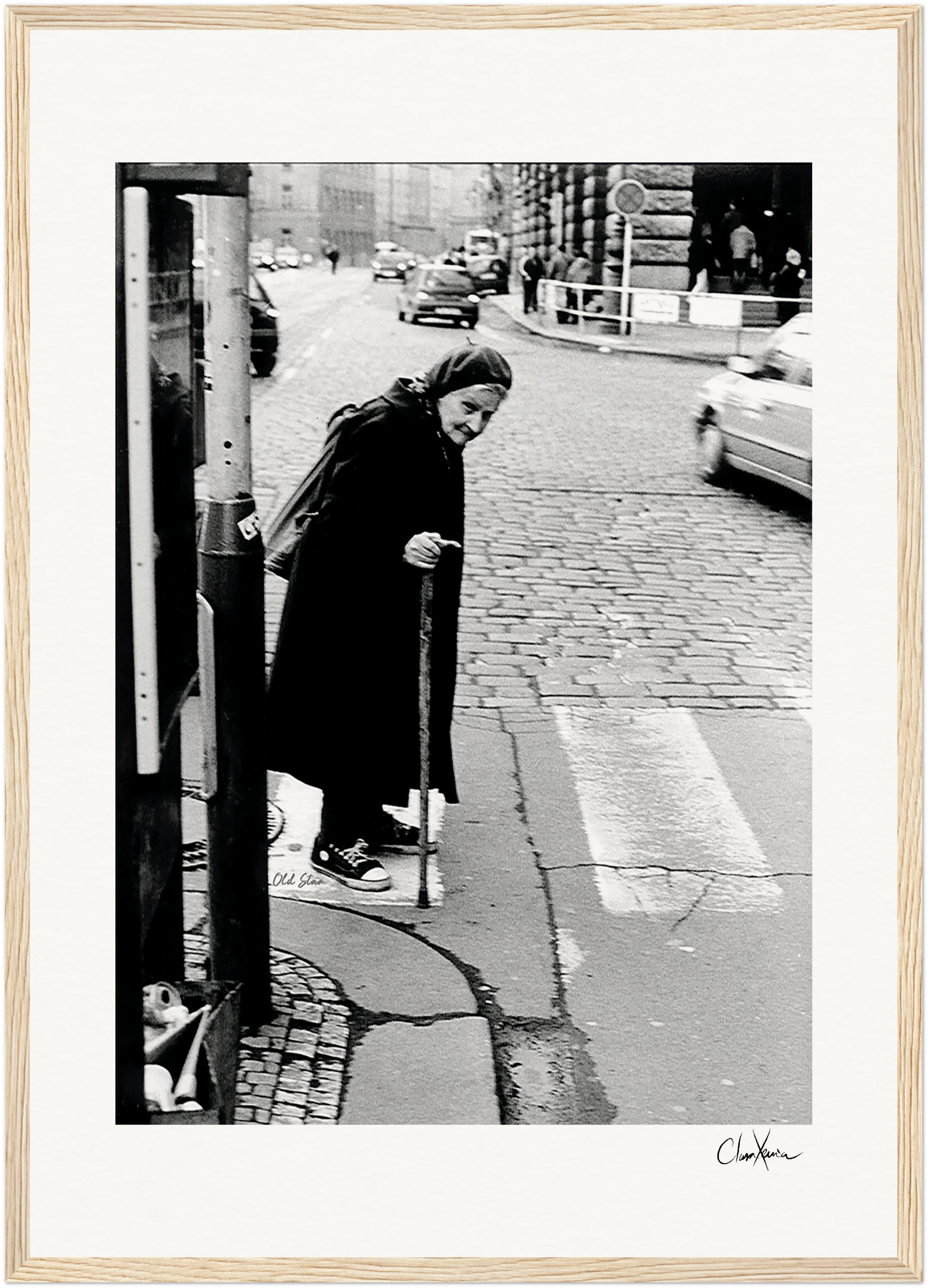 An elderly woman with a headscarf and cane stands at the edge of a cobblestone street, ready to cross. Cars and people appear in the background. This black-and-white image is framed in light wood, perfect as inspirational wall art for conscious interior design.