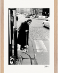 An elderly woman with a cane stands at the edge of a cobblestone street, preparing to cross. Cars and buildings line the background, and a payphone is visible in the foreground. This gallery-quality frame highlights the fine art print beautifully.