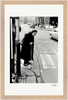 An elderly woman with a cane stands at the edge of a cobblestone street, preparing to cross. Cars and buildings line the background, and a payphone is visible in the foreground. This gallery-quality frame highlights the fine art print beautifully.