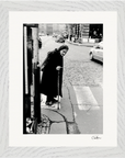 An elderly woman with a headscarf and cane stands at a street corner, preparing to cross a cobblestone road as cars pass by. This black-and-white fine art print is elegantly presented with a white border, perfect for conscious interior design.