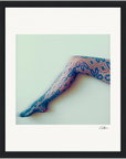 A single outstretched leg in patterned lace tights with a floral design is centered against a pale background. This fine art print, framed with a black border, adds elegance to any conscious interior design.