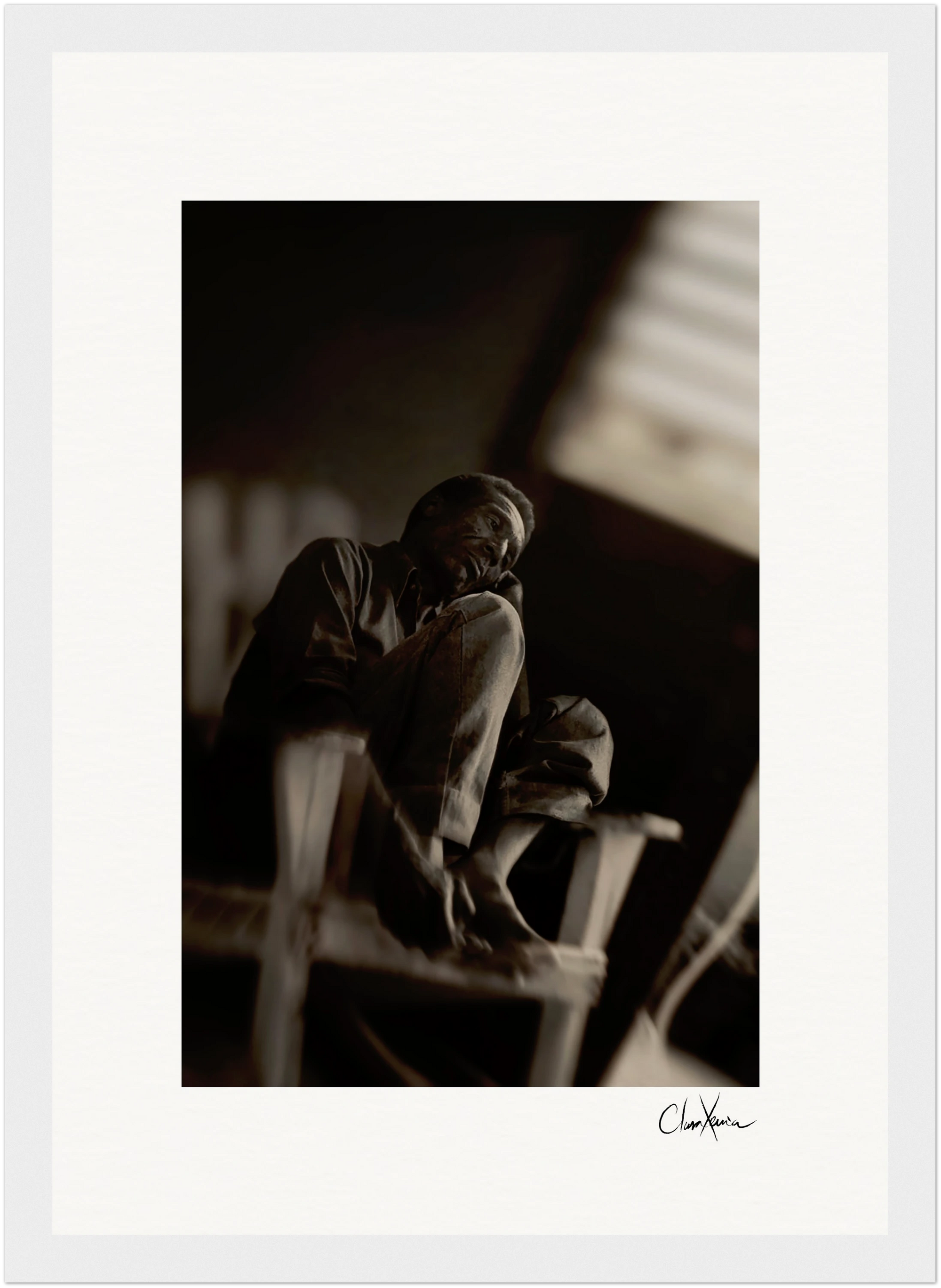 A man sits barefoot on a wooden chair with his knee pulled to his chest, gazing off to the side in a dimly lit room. This black and white fine art print features an artist’s signature at the bottom, perfect for inspirational wall art displays.