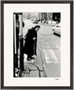 An elderly woman in a headscarf and long coat uses a cane to cross a cobblestone street at a crosswalk. Cars and buildings form the backdrop in this black-and-white fine art print, perfect for mindful home decor.