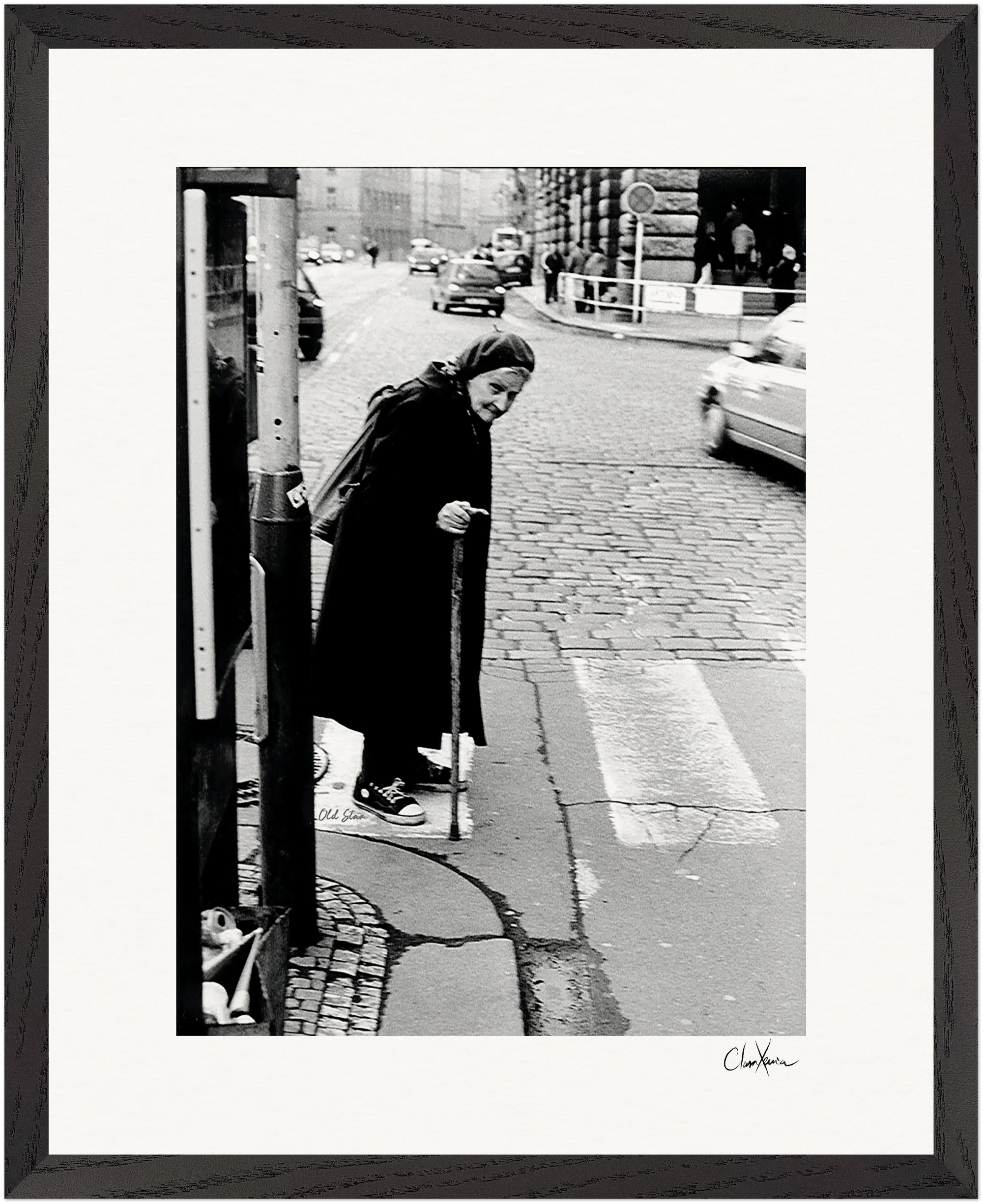 An elderly woman in a headscarf and long coat uses a cane to cross a cobblestone street at a crosswalk. Cars and buildings form the backdrop in this black-and-white fine art print, perfect for mindful home decor.