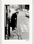 An elderly woman with a cane stands at a crosswalk on a city street, wearing a headscarf and long coat. This fine art print captures an inspiring moment for conscious interior design or inspirational wall art. Cars line the background.