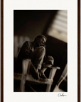 A black and white fine art print of a person sitting on a chair with knees up, gazing thoughtfully to the side. The dark, blurred background enhances its elegance. Framed with a white mat and dark wood frame; signed at the bottom right.