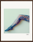 A single leg is extended against a light background, wearing sheer, blue lace tights with a floral pattern. With soft focus and a dark brown border, this fine art print enhances mindful home decor.