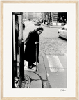 An elderly woman with a cane crosses a cobblestone street at a crosswalk, wearing a long coat and headscarf. Cars and buildings line the background in this black and white image—perfect as framed wall art for mindful home decor.
