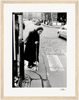 An elderly woman with a cane crosses a cobblestone street at a crosswalk, wearing a long coat and headscarf. Cars and buildings line the background in this black and white image—perfect as framed wall art for mindful home decor.