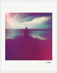 A silhouetted figure stands on a beach under a cloudy sky, with light flares and grainy effects creating a soft, dreamy look. This inspirational wall art features a white border and a signature in the corner—perfect for any gallery-quality frame.