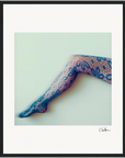 A single outstretched leg in blue floral lace-patterned tights against a pale background, highlighted as a fine art print with a black signature in the bottom right corner.