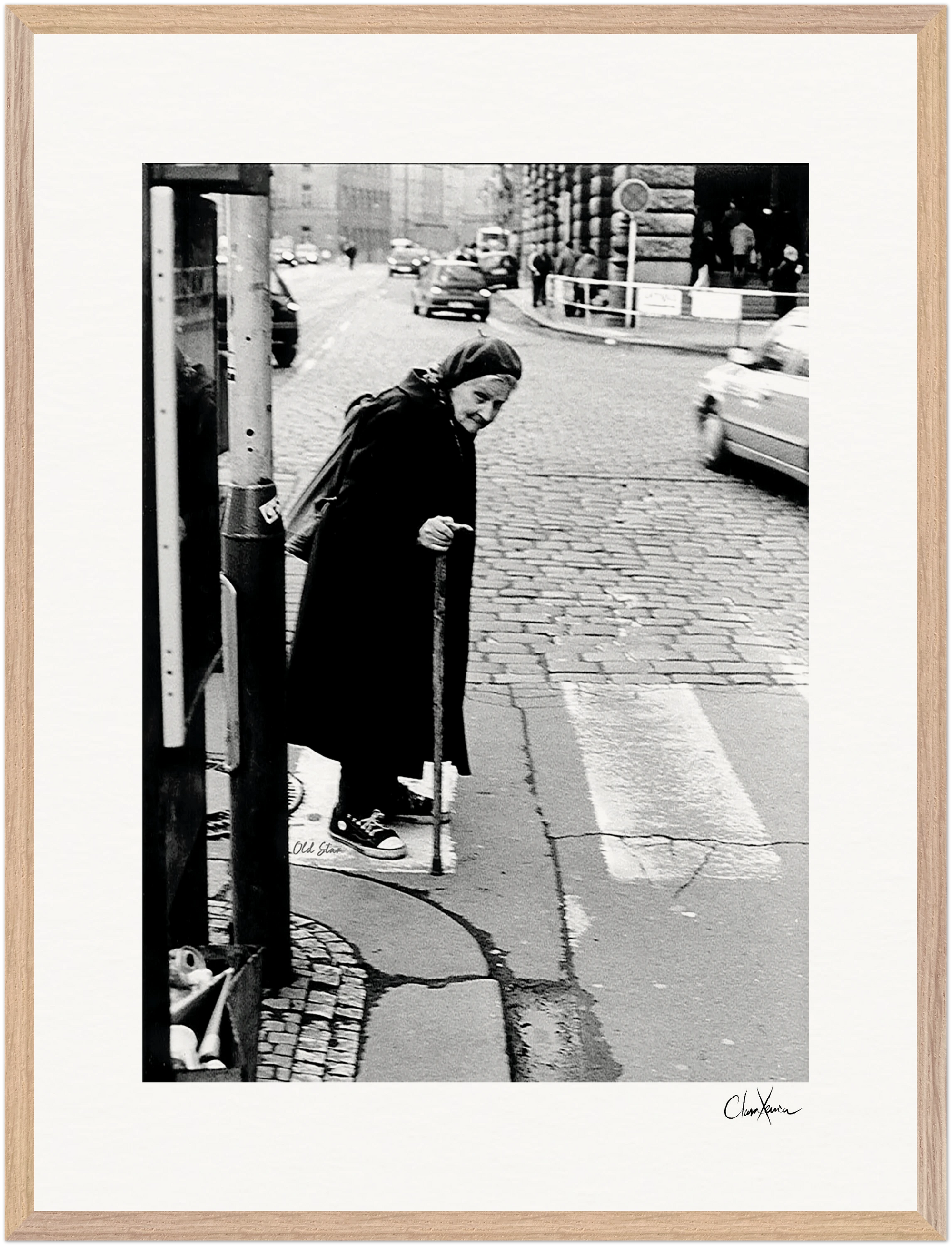 An elderly woman in a long coat and headscarf uses a cane to step off a curb at a crosswalk. Cars and buildings frame the scene. This black-and-white photo makes an elegant fine art print for mindful home decor.