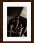 A black and white photo of a person sitting on a chair with legs crossed, resting their head on their hand, looking contemplative; framed in a gallery-quality frame with a wood texture and signed in the bottom right—perfect for mindful home decor.
