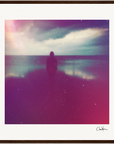 A silhouette of a person stands facing the ocean under a cloudy sky, with soft, colorful light and reflections on the water; this inspirational wall art has a hazy, dreamy effect and is beautifully framed with a brown wooden border.