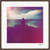A silhouette of a person stands facing the ocean under a cloudy sky, with soft, colorful light and reflections on the water; this inspirational wall art has a hazy, dreamy effect and is beautifully framed with a brown wooden border.