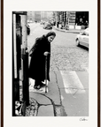 An elderly woman in a headscarf and dark clothing walks with a cane across a cobblestone street at a crosswalk, observed by cars and buildings—an evocative scene perfect for inspirational wall art or framed wall art in a mindful home decor setting.