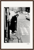 An elderly woman in a headscarf and dark clothing walks with a cane across a cobblestone street at a crosswalk, observed by cars and buildings—an evocative scene perfect for inspirational wall art or framed wall art in a mindful home decor setting.