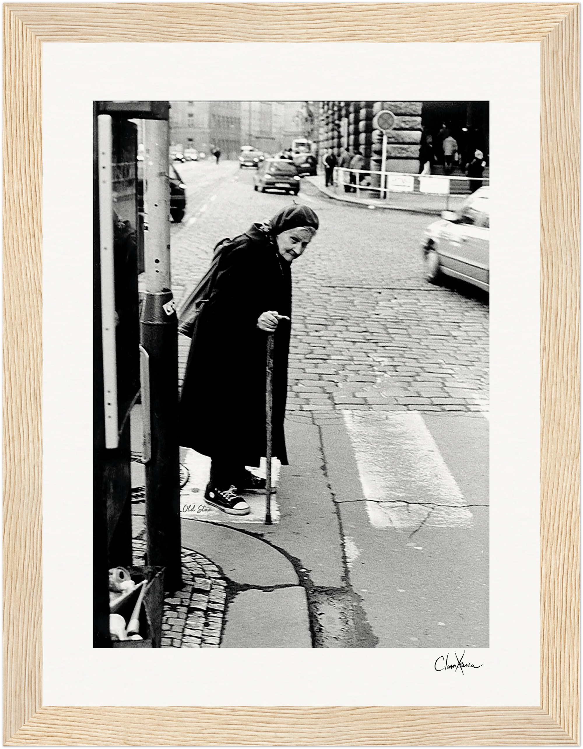 An elderly woman in a long coat and headscarf walks with a cane at a crosswalk on a cobblestone street. This black and white fine art print is elegantly framed with light wood, perfect for conscious interior design.