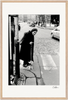 An elderly woman in a long coat and headscarf uses a cane to cross a cobblestone street at a crosswalk. Cars and people in the background evoke city life—perfect as framed wall art for mindful home decor.