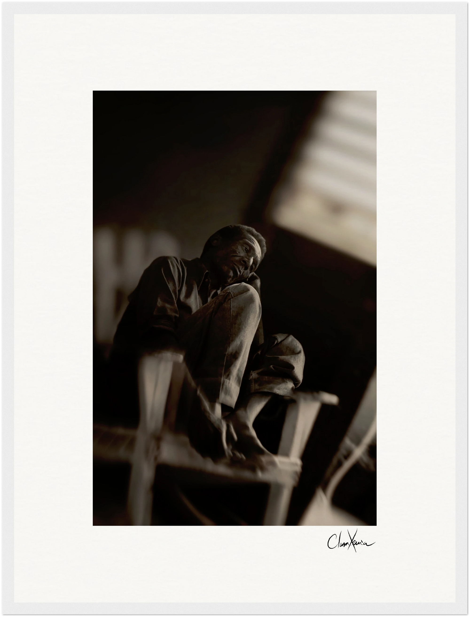 A man sits barefoot on a wooden chair, his arms resting on his knees, gazing thoughtfully into the distance. In black and white with soft focus and contrasted lighting, this fine art print is perfect as framed wall art for any conscious interior design.