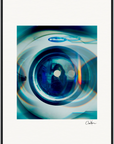 A close-up, slightly distorted image of the front door of an Electrolux washing machine, with a blue-green tint and circular light reflections. Perfect as a fine art print for mindful home decor, the artist's signature appears in the lower right corner.